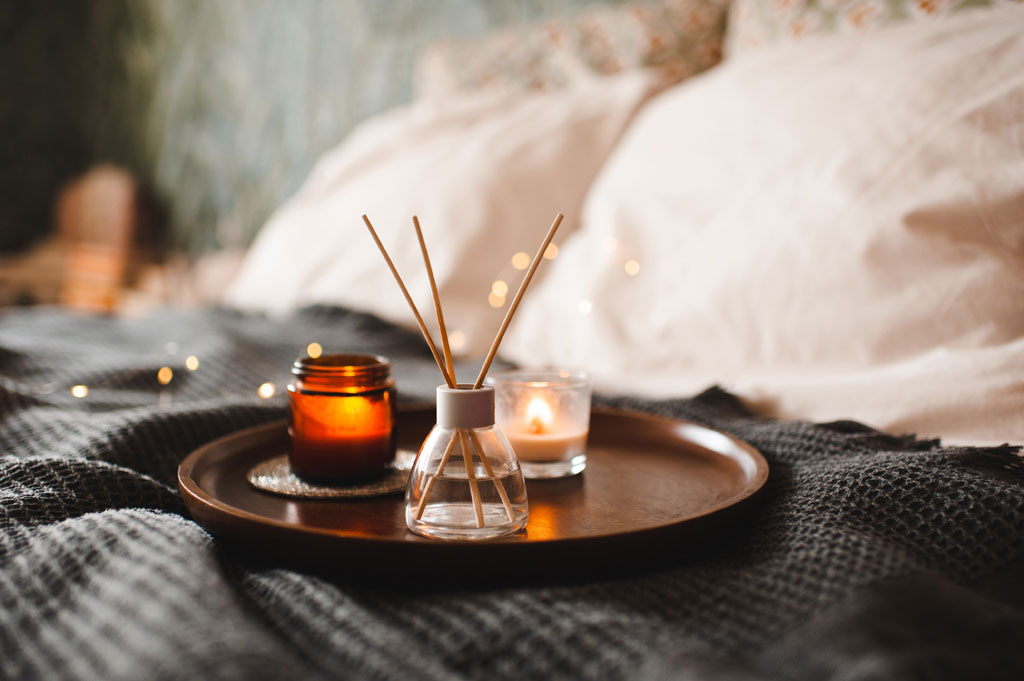 Fragrance diffuser with reed sticks and scented candles arranged on a tray in a cozy bedroom.